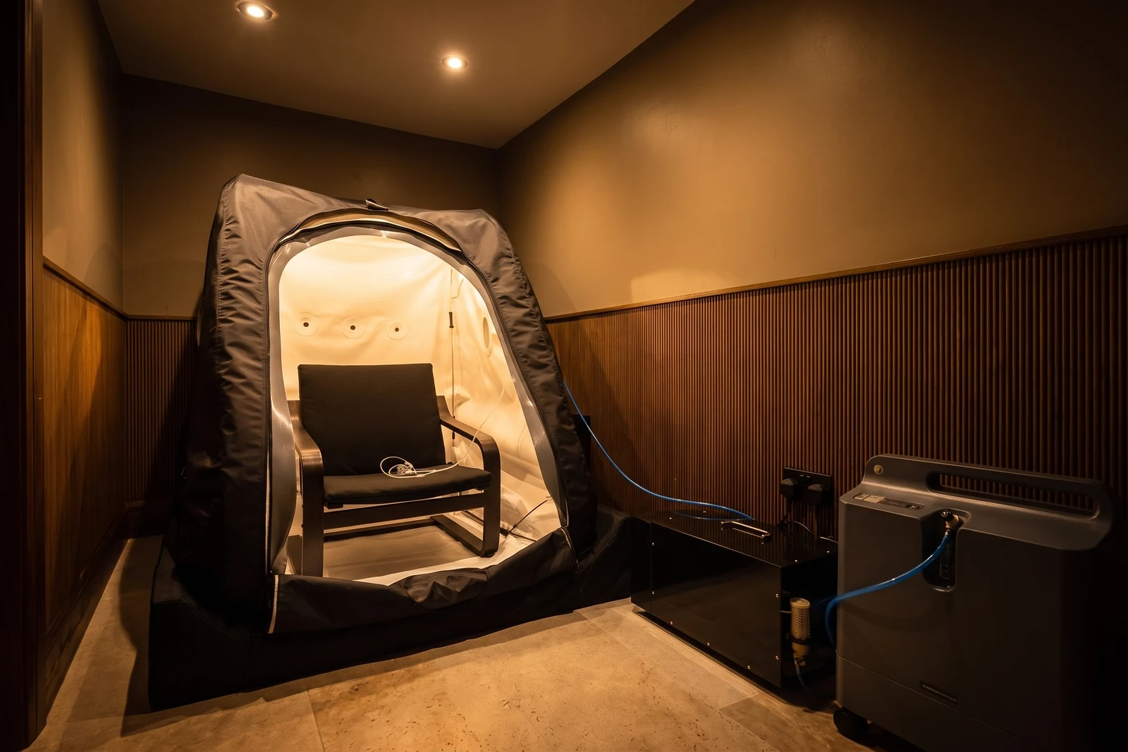 The Health Pod hyperbaric oxygen therapy room, with the chamber's arched doorway open and interior lit, beside an oxygen concentrator against timber-panelled walls.