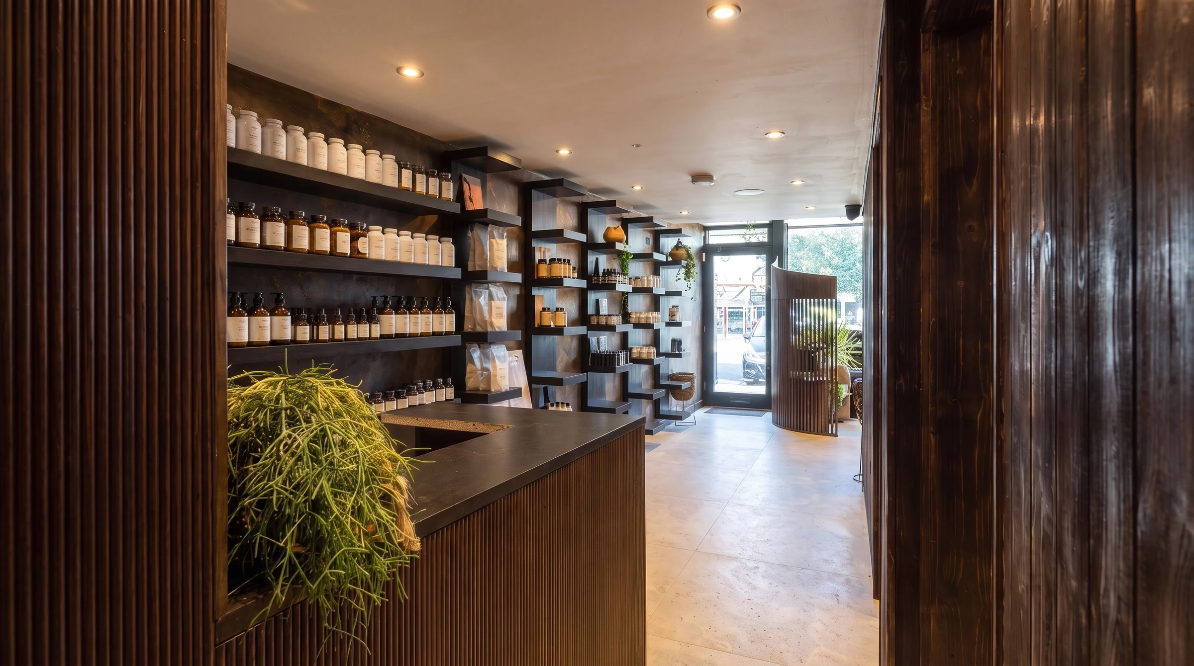 The Health Pod reception desk, a fluted-timber counter with a dark stone top and a trailing plant, beside black shelves of supplement jars and bottles, with the entrance and street beyond.