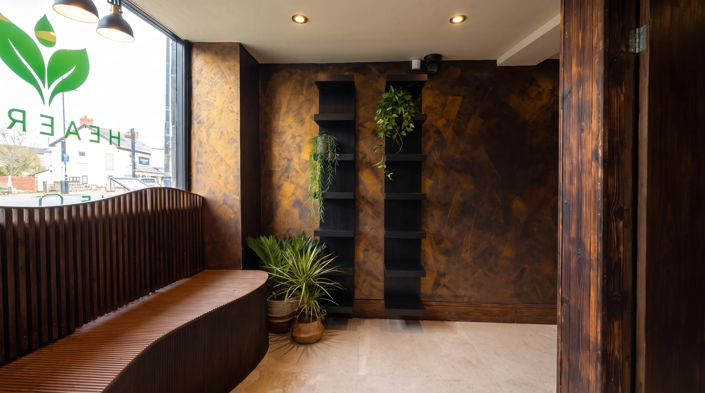 A quiet corner of the Health Pod studio, with a curved fluted-timber bench beneath the window, two black shelf columns holding trailing plants, and potted palms against a bronze plaster wall.